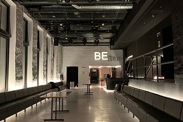 Blick an das Foyer des BE: Links und rechts entlang der Wände stehen schwarze Bänke, in der Mitte Tische. Am Ende des Ganges steht in Leuchtbuchstaben "BE".